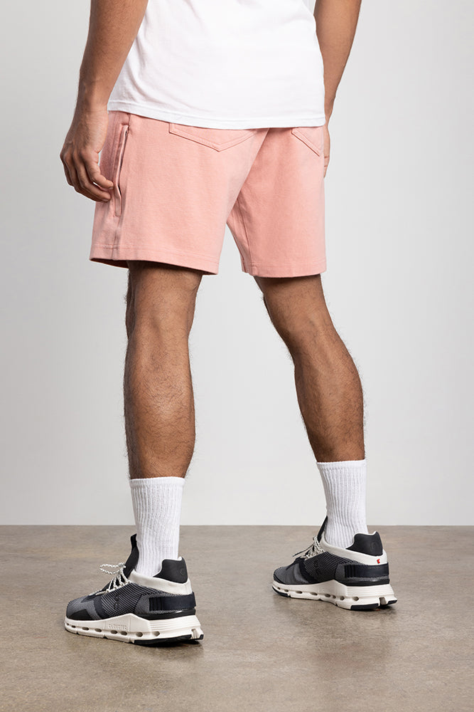 A person stands indoors on a concrete floor, seen from behind wearing a white t-shirt, 686 Mens Everywhere Double Knit Short in light pink, white crew socks, and black and white sneakers.