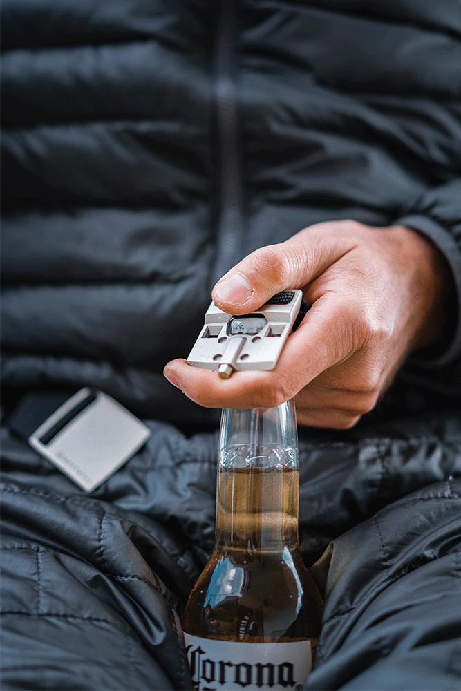 A person wearing a dark puffy jacket uses the 686 Westwell Clicker Stretch Belt as a multitool to open a bottle of Corona beer.